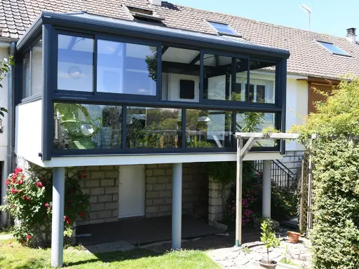 Une extension véranda toit plat moderne, avec de larges baies vitrées, s'intègre parfaitement à la maison. Un fauteuil vert est visible à l'intérieur.