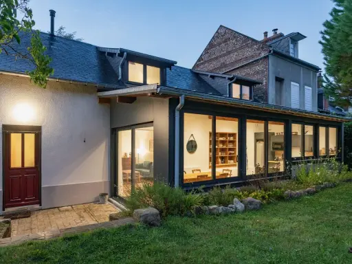 Extension maison en L avec grandes baies vitrées illuminant un salon et une cuisine. Une porte rouge s'ouvre sur une terrasse pavée.