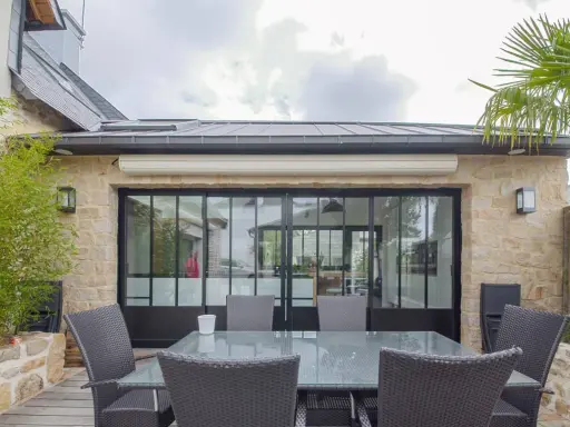 Terrasse avec mobilier autour d'une extension maison style atelier aux grandes verrières noires. Bambou et palmier apportent une touche végétale.