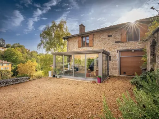 Maison en pierre avec une veranda lumineuse, un jardin de gravier et un paysage vallonné en automne.