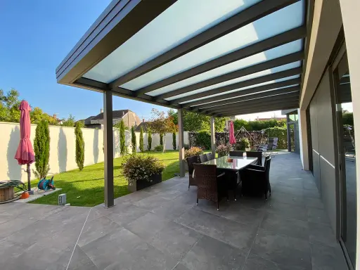Une belle pergola moderne avec un toit vitré pour couvrir l'espace repas extérieur. Le jardin est bien entretenu, avec une pelouse verte et des cyprès élancés.