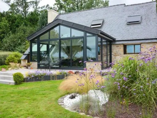 Maison avec extension toit vitrée, terrasse en bois et jardin fleuri.