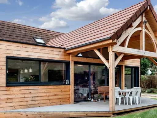 Extension de maison en bois avec terrasse sur dalle béton. Une table et des chaises sont installées sous un auvent.
