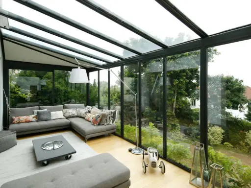 Salon lumineux avec canapé d'angle gris, table basse et vue sur jardin luxuriant. Idéal pour agrandir veranda et profiter de la nature.