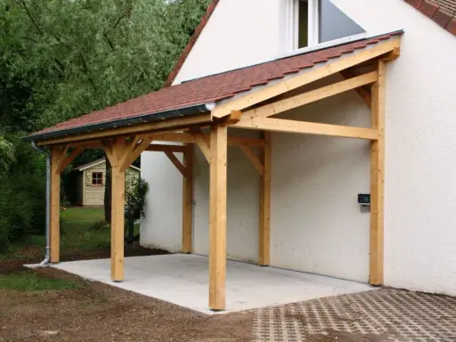 Un abri de voiture en bois avec une pente légère, adossé à une maison blanche.