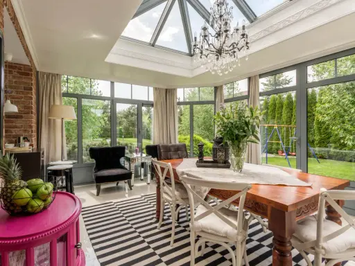 Une élégante veranda couloir avec un lustre, une table en bois et des fauteuils, donnant sur un jardin verdoyant.