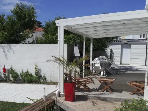 Terrasse en bois sous un préau semi-fermé, avec chaises empilées, une table, un palmier en pot et un barbecue.