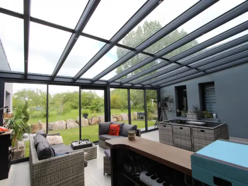 Salon moderne avec un toit en verre laissant entrer la lumière. Des canapés confortables invitent à la détente, tandis que le jardin verdoyant offre une vue paisible.