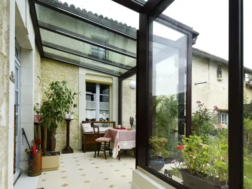 Une veranda avec toiture tuile, aménagée avec une table et des plantes, s'ouvre sur un jardin verdoyant.