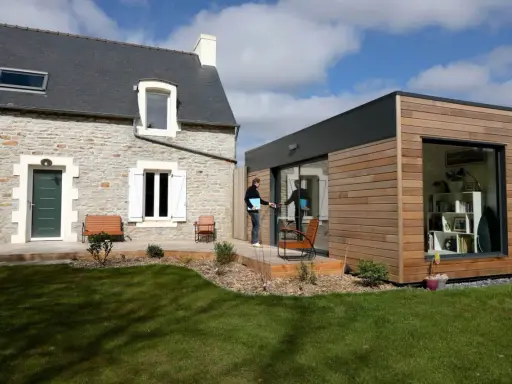 Une maison en pierre avec une extension moderne en bois. Un homme sort de l'extension, prêt à profiter du jardin.