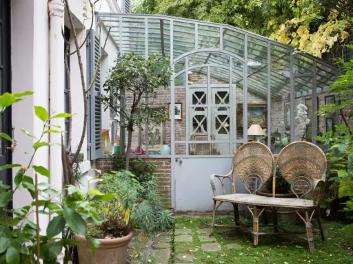 Une charmante veranda en fer forgé abrite un banc en rotin et des plantes luxuriantes, invitant à la détente dans ce jardin secret.