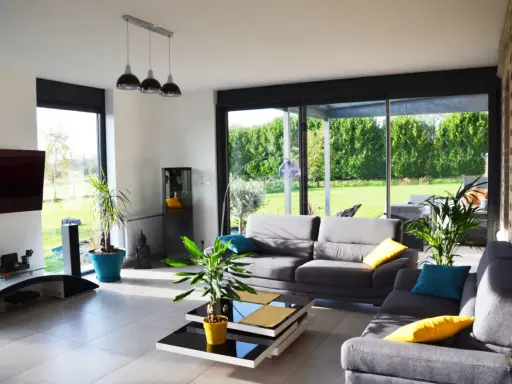 Salon lumineux avec canapés gris, table basse moderne et grandes baies vitrées donnant sur un jardin verdoyant. Une annexe maison parfaite pour se détendre.