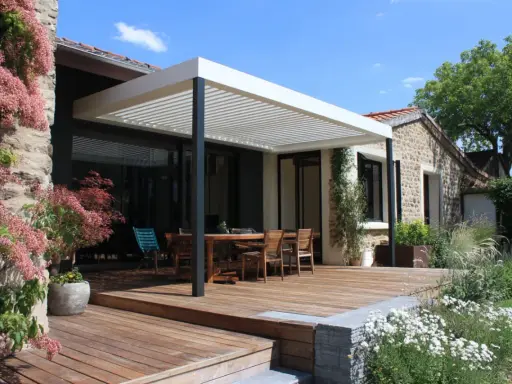 Terrasse en bois sous un toit pergola naturel, avec table et chaises, entourée de végétation luxuriante et d'une maison en pierre.