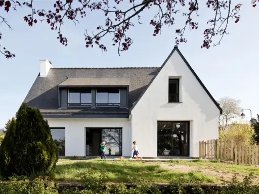 Une extension maison néo-bretonne moderne avec de grandes baies vitrées. Deux enfants jouent dans le jardin verdoyant devant la maison.