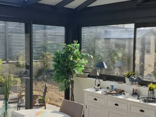 Intérieur lumineux d'une véranda triple vitrage, avec vue sur un jardin verdoyant et une commode blanche décorée.