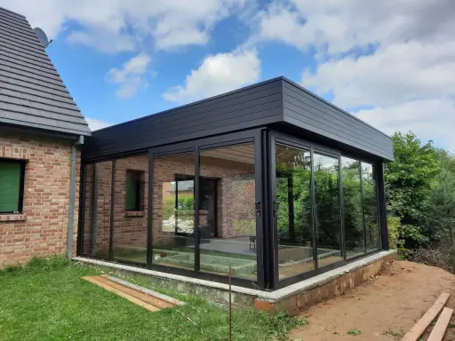 Moderne véranda toit plat avec puits de lumière, offrant une vue panoramique sur le jardin. L'extension en verre et métal s'intègre parfaitement à la maison en briques.