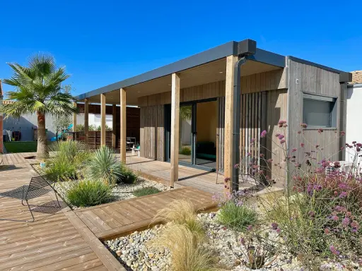 Maison moderne avec une extension toiture terrasse en bois, entourée d'un jardin paysager et d'une terrasse en bois.