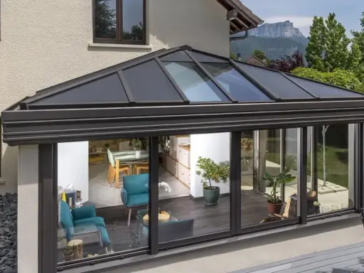 Une **pergola bioclimatique** moderne avec toit vitré, prolongeant une maison vers un jardin verdoyant. L'intérieur lumineux révèle un salon cosy et une salle à manger.