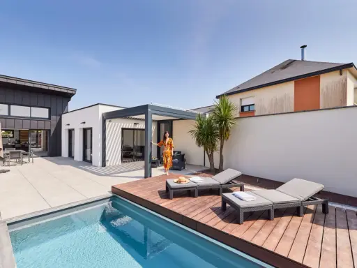 Une femme en robe colorée se tient près d'une pergola moderne, profitant de l'étanchéité pergola mur. Piscine et chaises longues complètent ce paysage de détente.