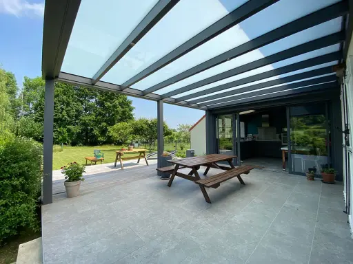Terrasse avec table en bois sous une pergola bioclimatique. La hauteur de la structure permet de profiter du jardin verdoyant.