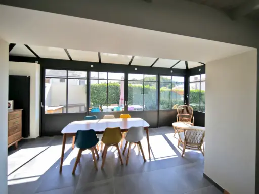 Salle à manger lumineuse dans une véranda chauffée, avec une table en bois et des chaises colorées. Un espace de vie agréable.