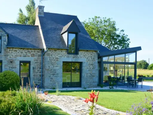 Maison en pierre avec une extension longere vitrée moderne. Un chemin pavé mène à la terrasse où une table est dressée.