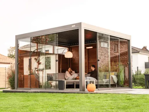 Un couple se détend dans une pergola moderne avec parois vitrées. La fixation pergola est discrète, laissant place à un espace cosy avec chauffage d'appoint.