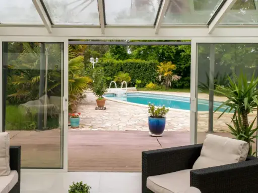Une extension vitrée moderne s'ouvre sur une piscine et un jardin luxuriant, invitant à la détente.
