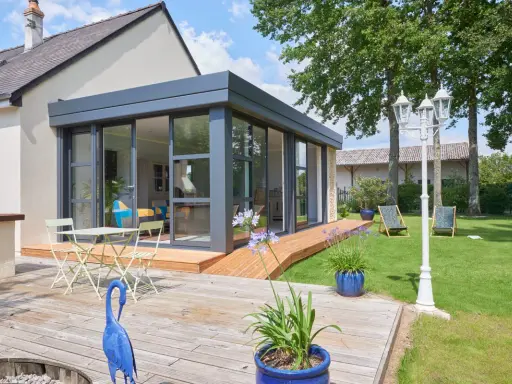 Extension toit plat sur maison ancienne, avec terrasse en bois, mobilier de jardin et vue sur un jardin verdoyant.