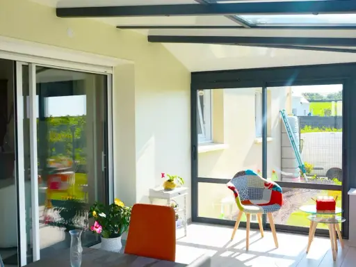 Intérieur lumineux d'une véranda avec un grand vitrage offrant une vue sur un jardin verdoyant. Des chaises colorées invitent à la détente.