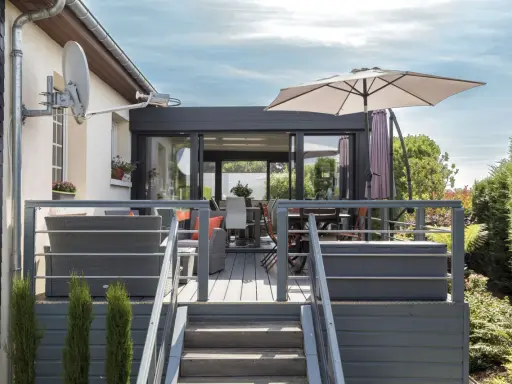 Terrasse avec mobilier de jardin et une véranda bois et alu. Parasol ouvert, escalier menant à la terrasse.