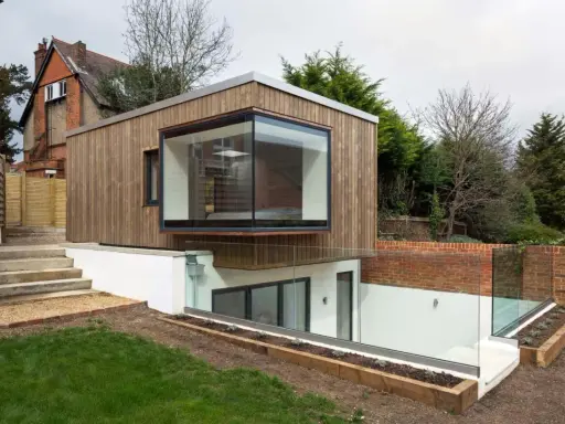 Une extension de maison moderne en bois avec une grande fenêtre en saillie. L'agrandissement maison s'intègre à un jardin en pente avec des murs de briques et des garde-corps en verre.