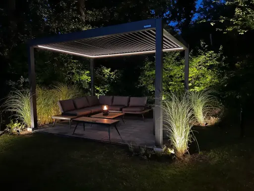 Une **pergola au milieu du jardin** illuminée la nuit, avec un salon de jardin confortable et des herbes hautes éclairées.