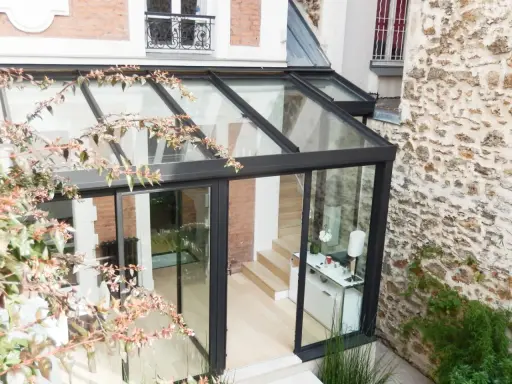 Une pergola transformée en véranda moderne, avec une structure en verre et métal, s'intégrant à une façade en pierre et brique.