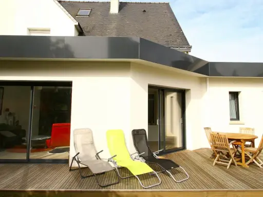 Extension de toit plat en bac acier, avec terrasse en bois, chaises longues et table de jardin.