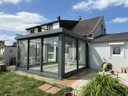 Une véranda moderne, conçue par un architecte, prolonge une maison. Elle est en verre et métal gris, avec un toit plat. Le jardin est verdoyant.