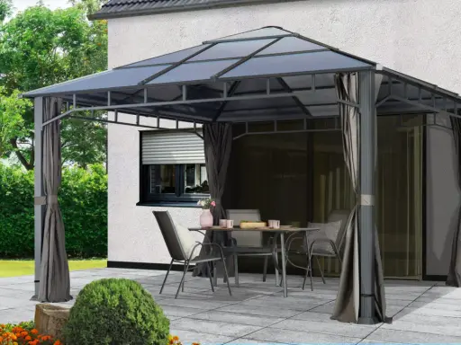 Une tonnelle def. avec table et chaises sur une terrasse pavée, devant une maison blanche et un jardin verdoyant.