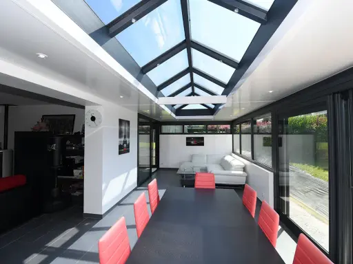 Une extension veranda salle à manger moderne avec un toit vitré, une table noire et des chaises rouges, un canapé blanc et une vue sur le jardin.