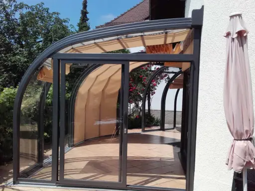 Une élégante veranda demi ronde en verre et métal, avec des stores rétractables, s'ouvre sur un jardin fleuri.