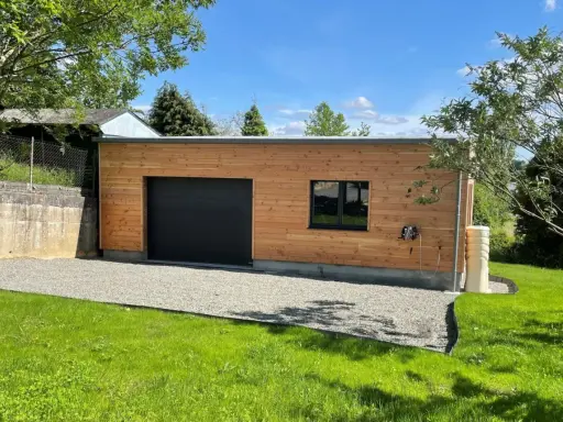 Extension garage bois accolé maison, avec porte noire, fenêtre et jardin verdoyant.