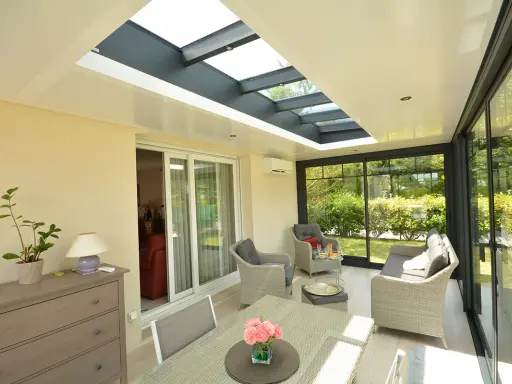 Une **veranda qui s'ouvre entièrement** sur un jardin verdoyant. Mobilier confortable, table et chaises pour profiter du soleil.