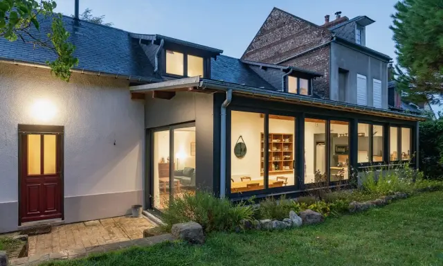 Extension maison en L avec grandes baies vitrées illuminant un salon et une cuisine. Une porte rouge s'ouvre sur une terrasse pavée.