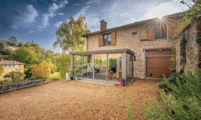 Maison en pierre avec une veranda lumineuse, un jardin de gravier et un paysage vallonné en automne.