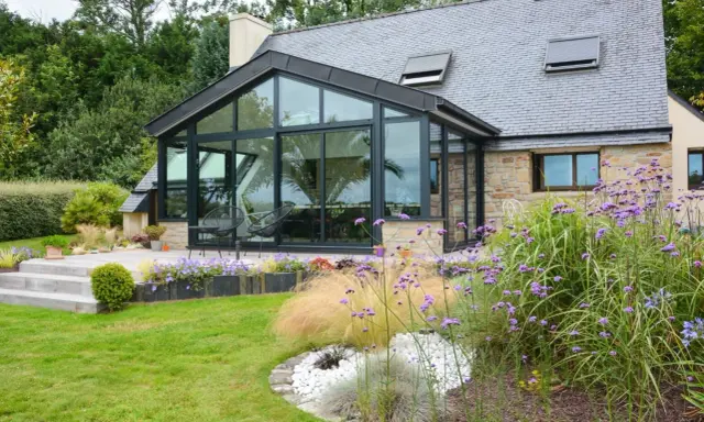 Maison avec extension toit vitrée, terrasse en bois et jardin fleuri.