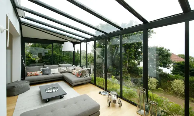 Salon lumineux avec canapé d'angle gris, table basse et vue sur jardin luxuriant. Idéal pour agrandir veranda et profiter de la nature.
