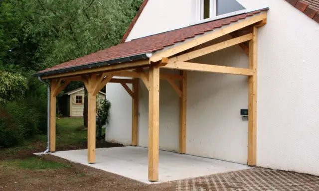 Un abri de voiture en bois avec une pente légère, adossé à une maison blanche.