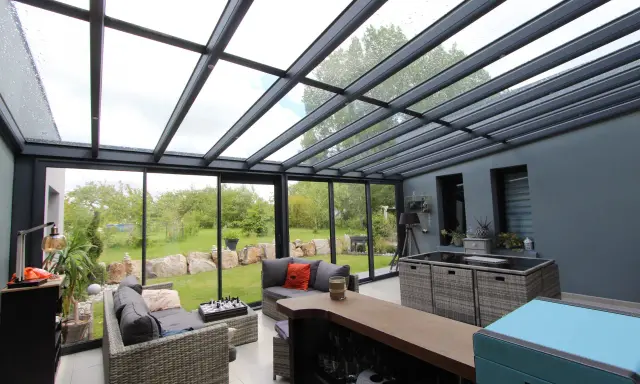 Salon moderne avec un toit en verre laissant entrer la lumière. Des canapés confortables invitent à la détente, tandis que le jardin verdoyant offre une vue paisible.