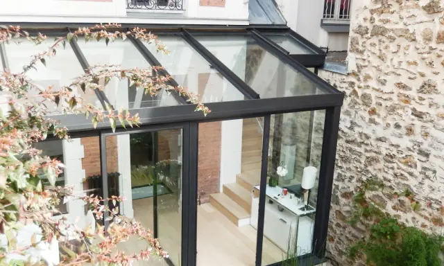Une véranda bioclimatique moderne, avec une structure en verre et métal noir, s'intègre à une maison en briques et pierre. Avis positif sur son esthétique.