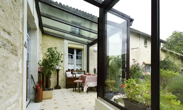Une veranda avec toiture tuile, aménagée avec une table et des plantes, s'ouvre sur un jardin verdoyant.