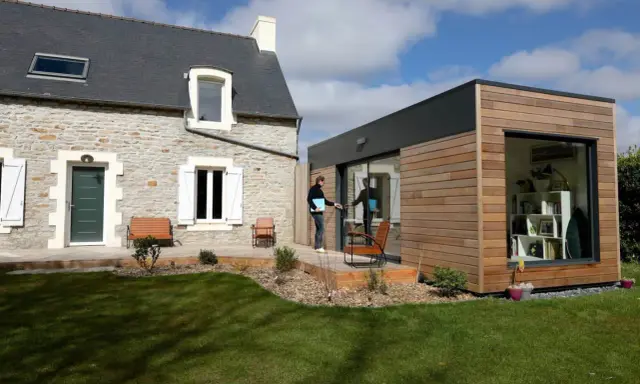 Une maison en pierre avec une extension moderne en bois. Un homme sort de l'extension, prêt à profiter du jardin.